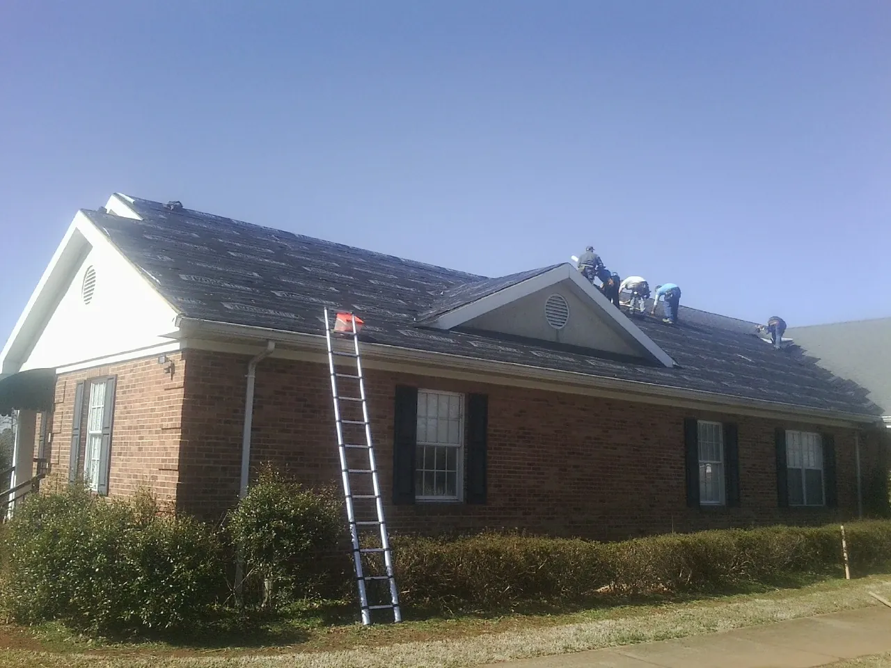 Crew tearing shingles off a brick ranch roof with a ladder set — Emergency Roof Repair work by our crew in Cornish
